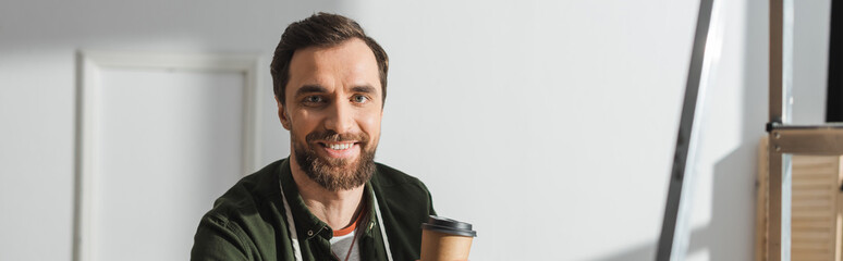 Positive bearded carpenter holding coffee to go and looking at camera in workshop, banner.
