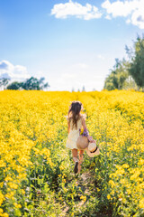 girl with flowers and a hat in her hand runs into a field of rapeseed. Yellow flower field. View from the back