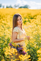 Naklejka na ściany i meble Portrait of a beautiful girl in yellow rapeseed flowers. A woman collects flowers in her purse