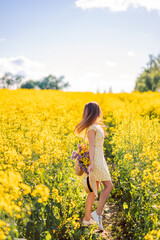 Fototapeta premium girl in a hat with a bag of flowers walks in a field of rapeseed. Yellow flower field. View from the back