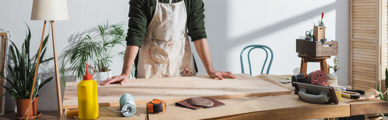 Cropped view of repairman in apron standing near wooden board and tools in workshop, banner. © LIGHTFIELD STUDIOS