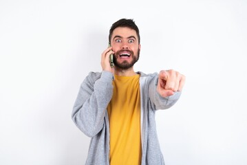 Fototapeta premium Positive caucasian man wearing casual sportswear over white wall indicates directly at camera has telephone conversation smiles broadly enjoys talking long hours. You join me