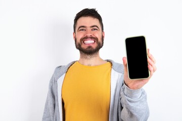 Charming adorable caucasian man wearing casual sportswear over white wall holding modern device, showing black screen smartphone