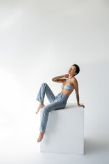 Obraz na płótnie Canvas young barefoot woman in blue jeans and satin bra posing on cube and looking at camera on grey background.