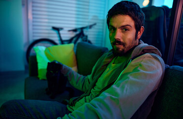 Man, portrait or neon lighting on sofa in living room, house or bachelor pad with feline touching, cat or domestic animal. Person, relax or pet kitten on couch in dark home, garage or night apartment