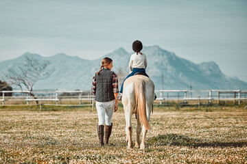 .Woman leading child on horse, ranch and mountain in background lady and animal walking on field from back. Countryside lifestyle, rural nature and farm animals, mom teaching kid to ride pony in USA.