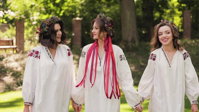 Three women walk holding hands and admire each other . Girls wear Ancient ethno Ukrainian embroidered shirt dress and wreath of flowers. Medium shot
