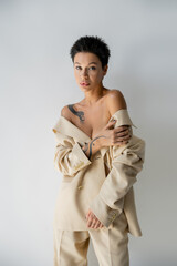 tattooed woman with naked shoulders posing in oversize suit while looking at camera isolated on...