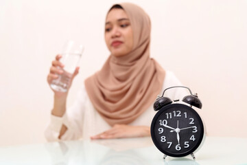 Young asian muslim woman taking a glass of water. Ramadhan concept. Selective focus on a clock