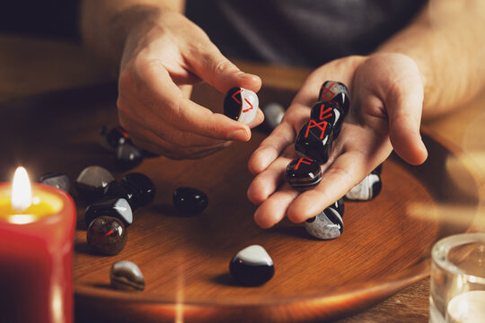 Man's hand holds a rune of jera with fingers. He shows runes on another hand. There are stones with different runes on a wooden tray. Red candle in foreground. Fortune telling. Close up