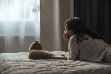 Sad young woman in pajamas with seasonal affective disorder lies alone on the bed near the window, next to the domestic cat. The concept of winter depression due to lack of sunlight
