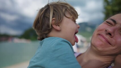 Funny cute moment of child kissing mother in cheek with tongue. Real life authentic family motherhood concept. Little boy bonding with parent outdoors - Powered by Adobe