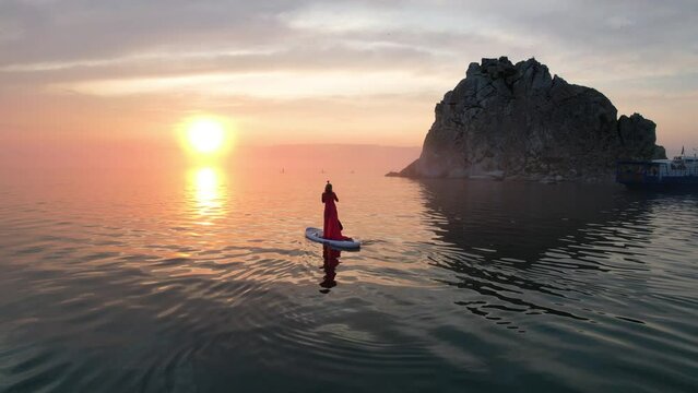 A girl in a red dress swims on a sup board at Shamanka Rock on Olkhon Island. Sunset on Lake Baikal in summer.