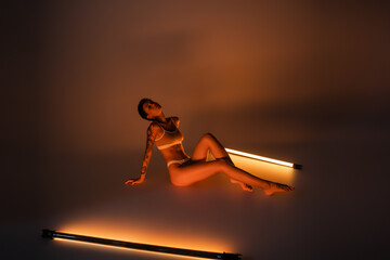 full length of seductive woman with slim tattooed body sitting in underwear near fluorescent lamps on dark background.
