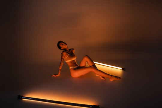 full length of seductive woman with slim tattooed body sitting in underwear near fluorescent lamps on dark background.