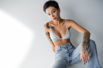 brunette woman with sexy tattooed body sitting in jeans and bra on grey background.