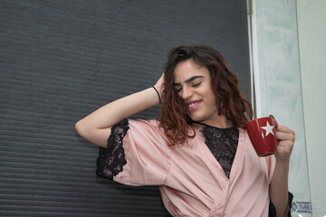 Young transgender girl, having a cup of coffee in the kitchen of her home in the morning, while she is stretching happy and smiling. Concept transgender, lgtbi, homosexuality, gay, diversity.
