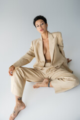 shirtless and barefoot woman in elegant pantsuit looking at camera while sitting on grey background.