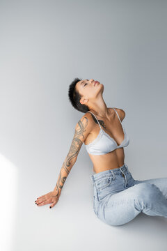 seductive tattooed woman in silk bra and jeans sitting with closed eyes on grey background.
