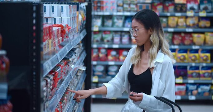 Young woman 20 teenager in glasses in grocery supermarket chooses groceries and consumer goods. Asian woman consumer with shopping cart in hand buys grocery in food supermarket. Shopping at food store