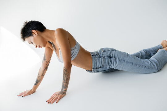 sensual tattooed woman in satin bra and blue jeans posing on grey background.