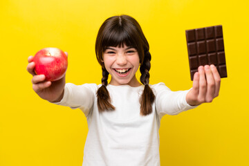 Little caucasian girl isolated on yellow background taking a chocolate tablet in one hand and an...