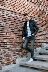 Fototapeta na wymiar A fashion male model on staircase. Young attractive caucasian man wearing black leather jacket and jeans. Young man wearing casual clothing on the street.