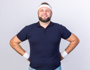 smiling adult slavic sporty man wearing headband and wristbands putting hands on waist and looking at camera isolated on white background with copy space