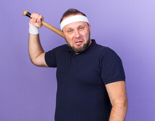 annoyed adult slavic sporty man wearing headband and wristbands holding bat on shoulder isolated on purple background with copy space