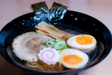 Soy sauce ramen in a black cup topped with chashu pork, Naruto fish balls, soy-sauce pickled eggs, menma bamboo shoots and seaweed. Ramen is a distinctive Japanese style noodle soup.