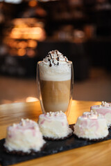 Latte with foam and cream in a transparent cup and cakes on a table in a cafe. Coffee with dessert to taste. Isolated image of dessert on bokeh background
