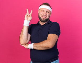 aching adult slavic sporty man wearing headband and wristbands gesturing victory sign isolated on pink background with copy space