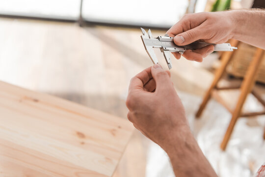 Cropped view of carpenter holding calipers near blurred wooden board in workshop.