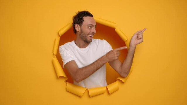 Horizontal shot of handsome caucasian man in white t-shirt points index finger right shows some promotion smiles gladly poses through paper hole