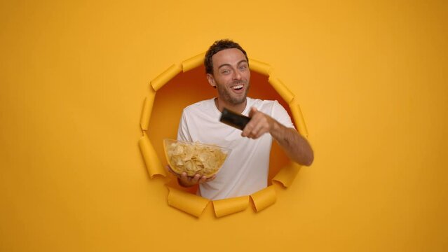 Indoor shot of focused brunette man holds bowl of chips and remote control chooses tv program looks concentrated wears casual t-shirt and sleeping mask poses through torn paper hole