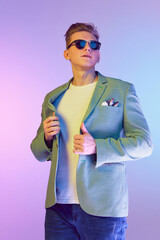 Modern stylish look. Young handsome man in stylish jacket and trendy sunglasses posing over purple background in neon light. Concept of emotions, lifestyle, youth, modern fashion. Ad