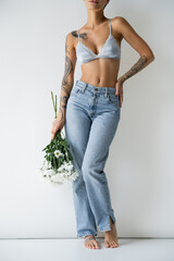 partial view of tattooed barefoot woman in silk bra and jeans standing with hand on hip and white...