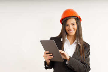 Portrait of young woman with tablet in her hands, dressed in business clothes on isolated white background. Studio shot of attractive happy brunette girl in orange construction helmet on her head.