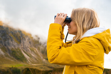 Side view of traveling girl looking through binoculars in mountains covered by clouds in a sunny day