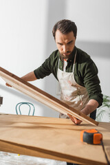 Fototapeta na wymiar Bearded craftsman in apron holding wooden board near ruler in workshop.