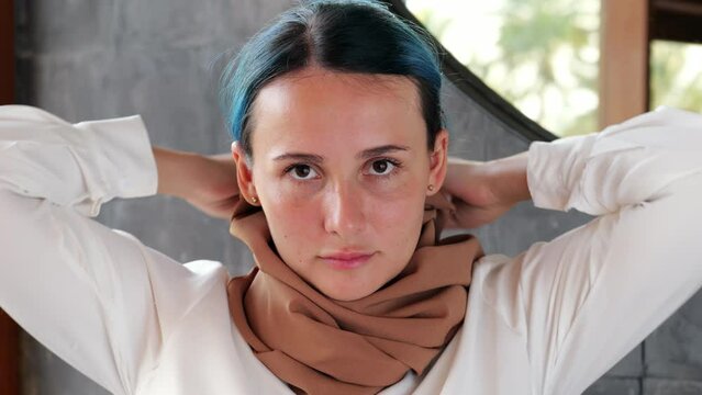 Young woman wearing a headscarf looks directly into the camera and pulls down her hijab, exposing her hair. Close indoor portrait shot with blurred background