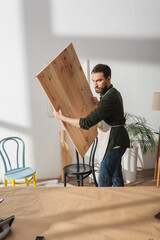 Carpenter holding wooden board near chairs in workshop.