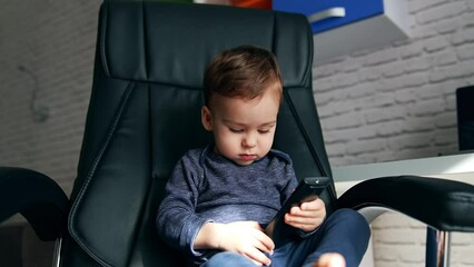 Serious little toddler sitting in a black leather chair. Beautiful child turning remote control in hands and pressing buttons.