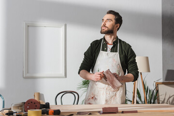 Bearded craftsman in apron holding sandpaper near blurred wooden board on table.