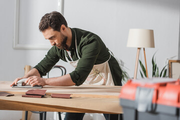 Repairman in apron sanding wooden plank near blurred toolbox in workshop.