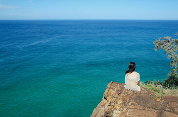 Back view of one young adult female sitting high up on a rock looking out to turquoise blue ocean and the horizon, with space for text or copy