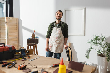 Smiling craftsman in apron standing near blurred tools on table.