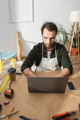 Bearded craftsman in apron using laptop near tools and coffee to go on table in workshop.