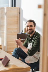 Smiling carpenter in apron looking at camera while sitting in workshop.