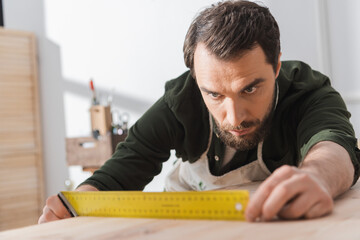 Focused carpenter looking at square tool on blurred wooden board.
